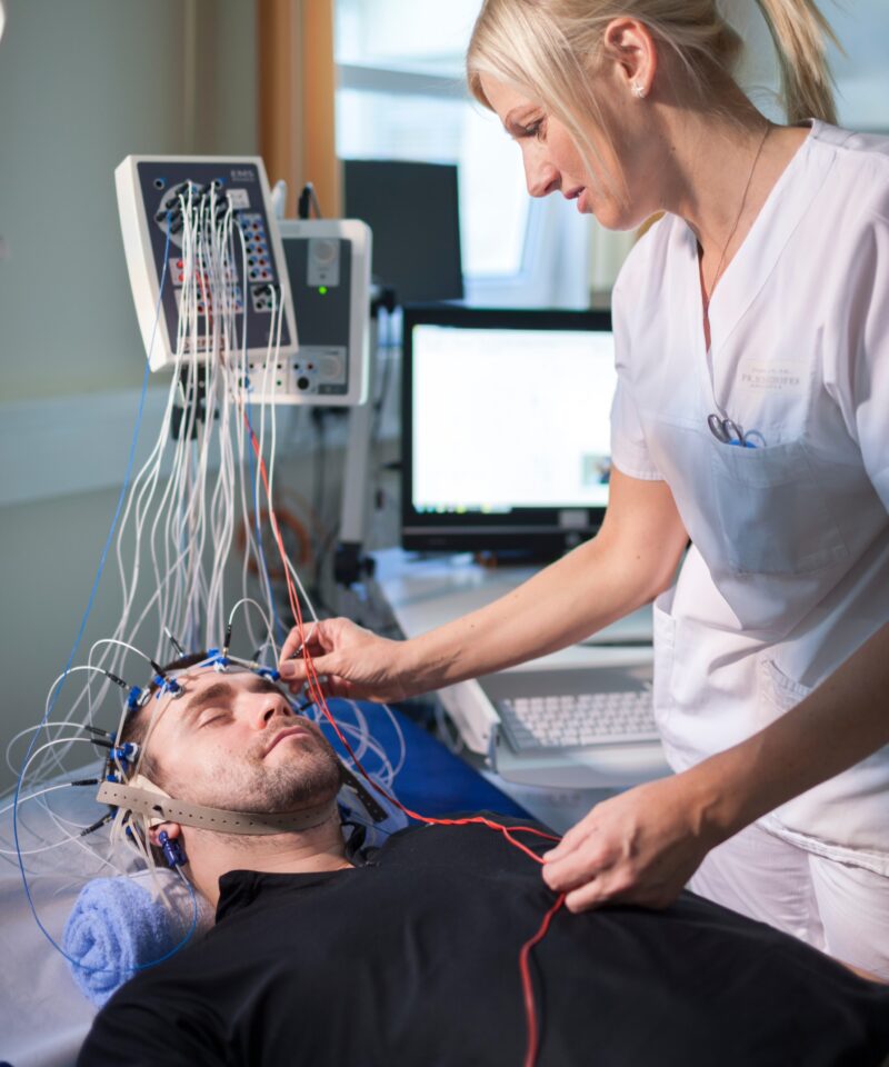 eine diplomierte Pflegekraft bringt gerade die Elektroden über eine Haube am Kopf am Patienten an