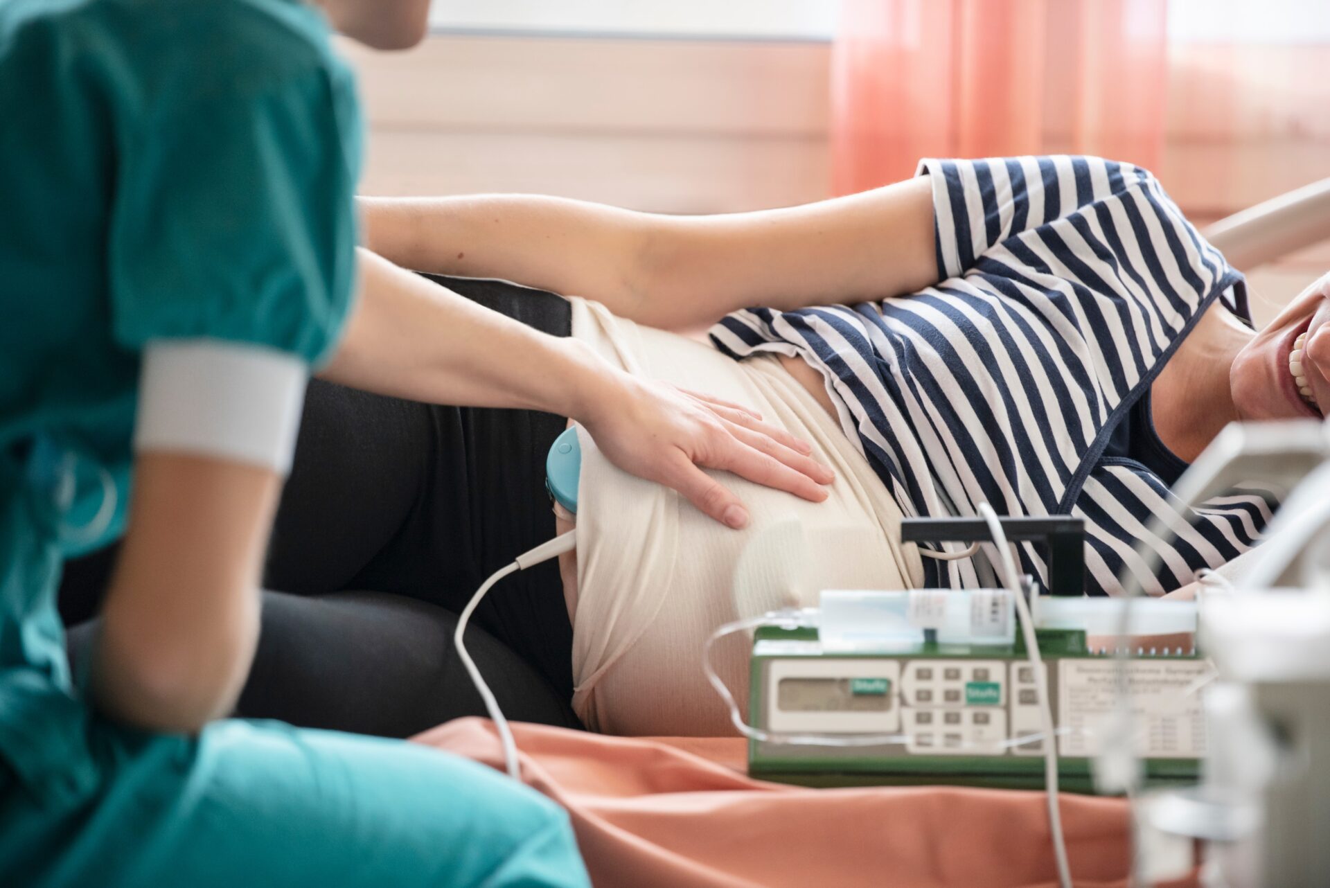 die Hebamme prüft die Herztöne des Babys mittels CTG (Kardiotokograf): Ein größerer Herzton-Wehenschreiber. Die Hebamme befestigt dafür zwei Sensoren mit Gurten am Bauch. Das Gerät zeichnet die Herztöne des Babys und eventuelle Wehen der Mutter gleichzeitig auf.