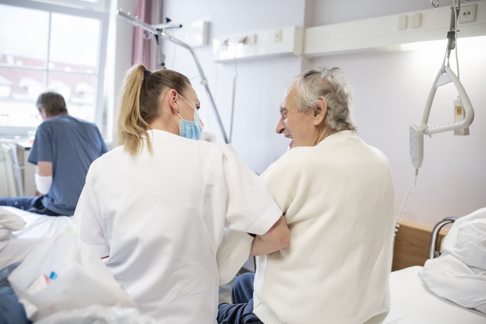 Die DGKP hilft einem älteren Patienten beim Aufstehen vom Patientenbett