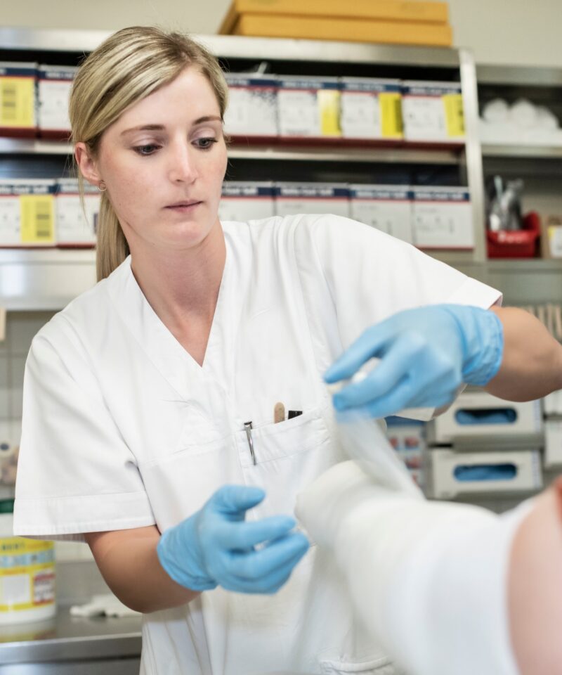 eine diplomierte Pflegekraft legt die feuchten Gips- oder Fiberglasbinden ohne Zug spiralförmig von unten nach oben gewickelt auf den Arm der Patientin