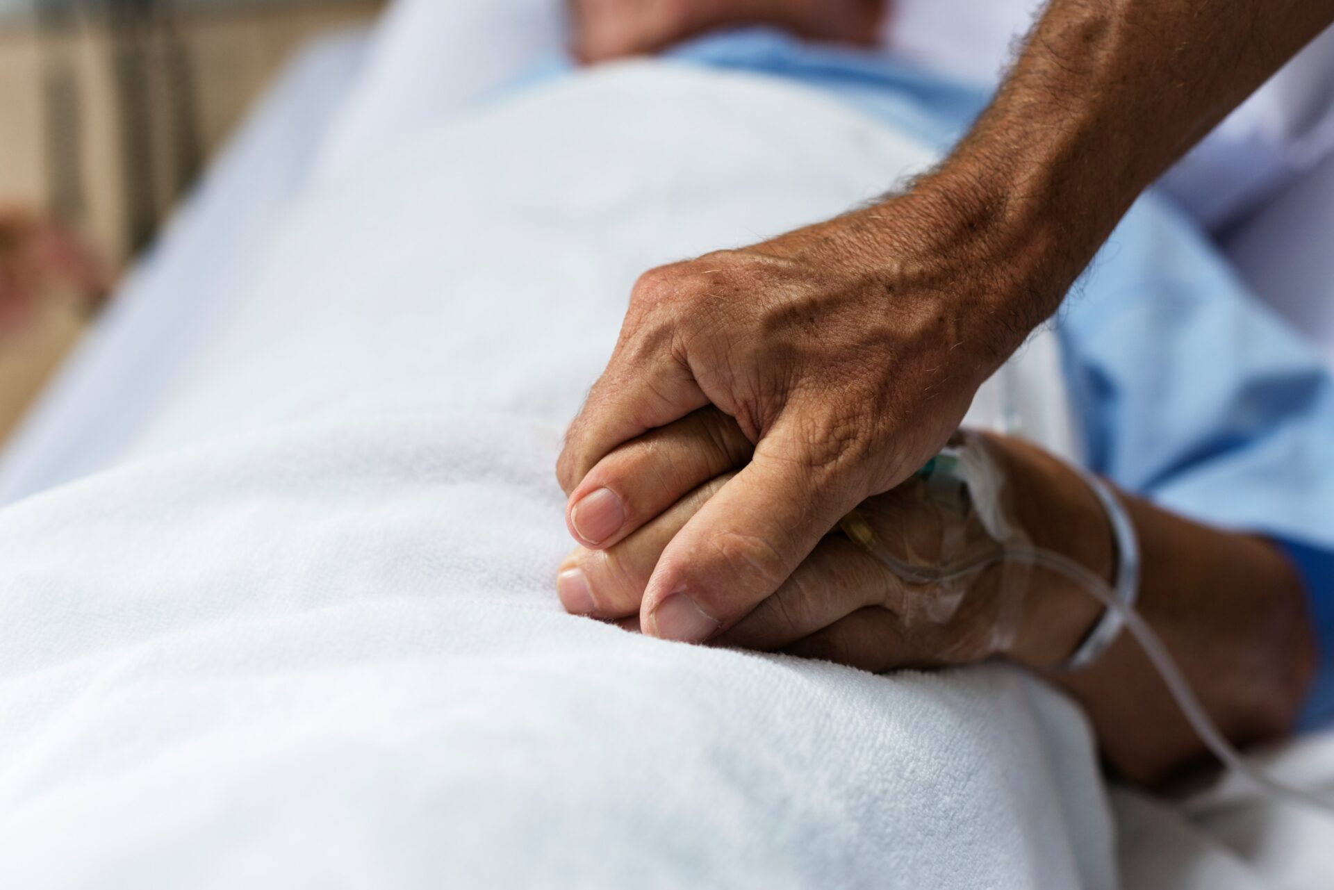 ein Angehöriger hält die Hand eines älteren Patienten, der im Patientenbett liegt